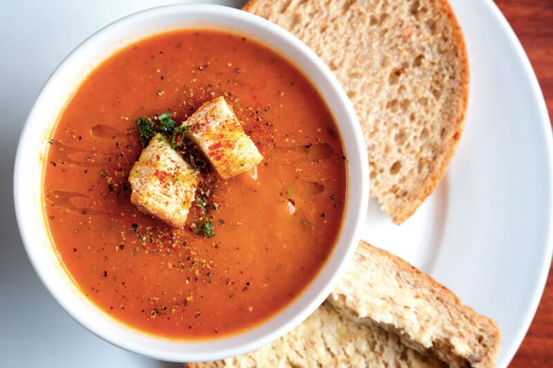 Tomatensoep met croutons in een witte kom, geserveerd met brood ernaast.