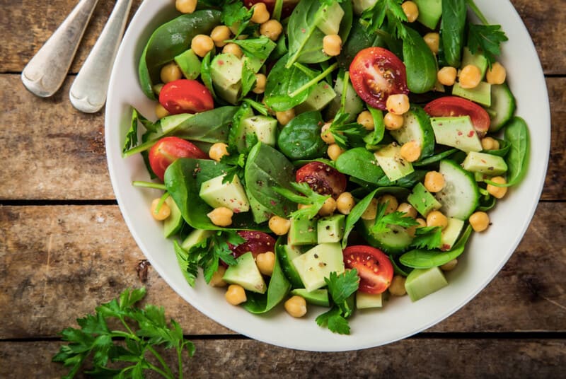 Salad with chickpeas, tomatoes, baby spinach, cucumber, avocado and parsley served in a white bowl. 