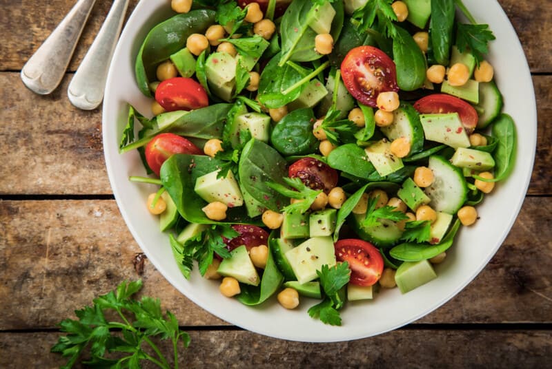 Salade met kikkererwten, tomaten, baby spinazie, komkommer, avocado en peterselie geserveerd in een witte kom.